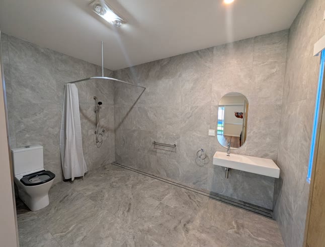 Roll-in wet-room ensuite with a level floor, linear drain, ceiling-track shower curtain, grab rail and a wall-hung basin with clear space beneath.